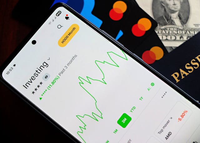 A smartphone with an investment graph on the screen atop credit cards, cash, and a passport.